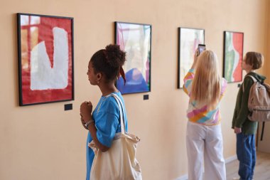 Sanat galerisi veya müzesinde soyut sanat eserlerine bakan çeşitli gençler ön planda Afro-Amerikan bir kızla aynı sırada duruyorlar.