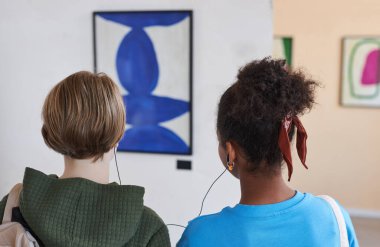 Modern sanat galerisinde veya müzede ses rehberiyle kulaklıkları paylaşan iki gencin arka plan görüntüsü