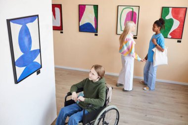 Engelli bir gencin yüksek açılı portresi sanat galerisindeki soyut resimlere bakıyor ve ses rehberini dinliyor, kopyalama alanını dinliyor.
