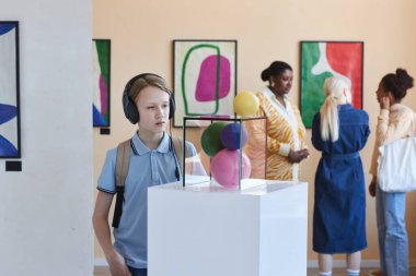 Kulaklık takan okul çocuğunun portresi müzede soyut sanat heykellerine bakıyor.