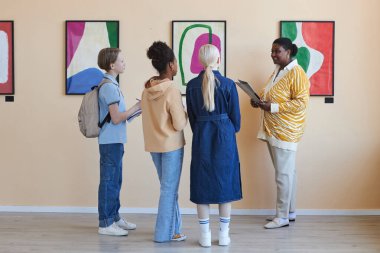 Çağdaş sanat galerisinde kadın öğretmenleri dinleyen çeşitli gençlerin renkli arka plan görüntüsü.