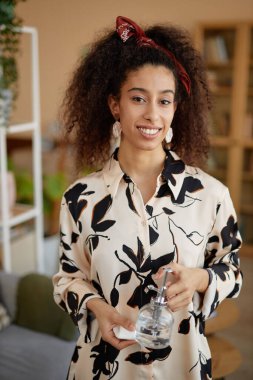 Kıvırcık saçlı genç siyahi bir kadının dikey portresi. Elinde küçük bir su kovası var ve kameraya gülümsüyor.