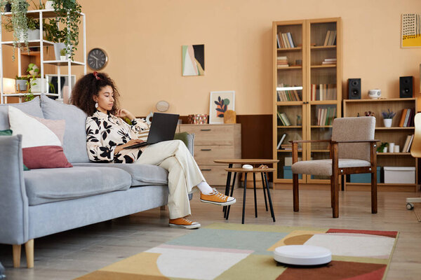 Wide angle view at young woman relaxing on couch with smart robot vacuum cleaner mopping floors copy space