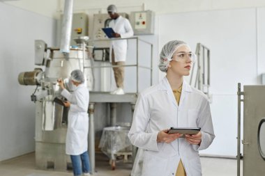 Laboratuvar önlüğü giyen ve ilaç fabrikasının temiz atölyesinde tablet tutan genç bir kadının belini kaldırın.