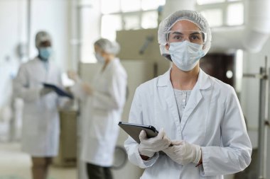 Maske takan ve laboratuvar önlüğü giyen genç bir kadının belden yukarı portresi. Fabrika atölyesinde kameraya bakıyor.