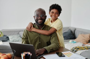 Kameraya gülümseyen ve dizüstü bilgisayarla çalışırken babasına sarılan mutlu bir çocuğun portresi.