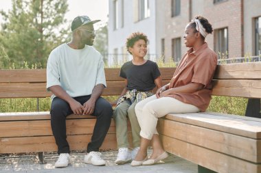 Afro-Amerikalı bir aile çocuğuyla birlikte bankta oturuyor ve birbirleriyle konuşuyorlar.