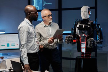 Çok ırklı iki mühendis ofiste dururken dijital tablet kullanan robotun programını inceliyor.