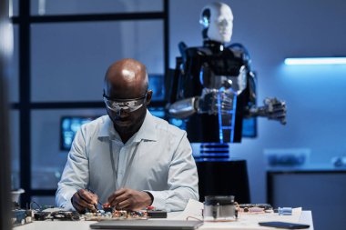 Laboratuvarda laboratuvar önlüğü giyen Afrikalı Amerikalı mühendis lehimleme masasında anakart kullanıyor.