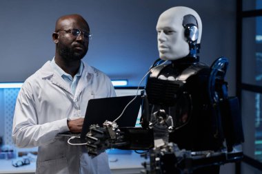 Laboratuvar önlüklü Afrikalı Amerikalı mühendis ofisindeki robotu kontrol etmek için laptop kullanıyor.