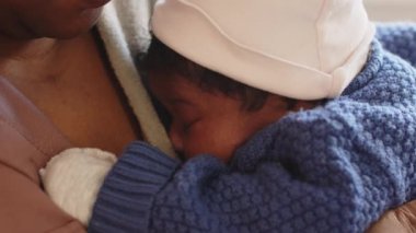 Uykulu, Afro-Amerikan bebek, mavi örgü şort ve beyaz kazı kazan eldivenleri giymiş, annesinin kollarında uykusundan uyanan.