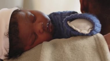 Beyaz bebek şapkası takmış barışçıl Afrikalı Amerikalı bebeğin orta boy yakın çekimi, eldivenler çizilmiş ve evde annesinin göğsünde uyuyan mavi bir tulum örülmüş.