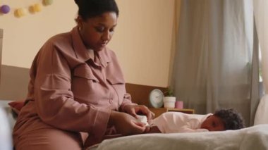 Genç siyahi bir kadının rahat bir dairede yatıp, küçük bebeğine sırt masajı yaptığı orta boy bir fotoğraf.