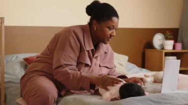 Orta boy Afro-Amerikalı bir kadın, küçük bebeğini kucağında kucağında dizüstü bilgisayarla rahat bir yatak odasında otururken okşuyor.