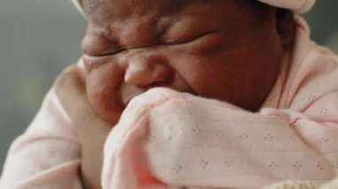 Eldivenleri annesinin kollarında ağlayan, bebek şapkası takmış, açık pembe tulumlu Afro-Amerikan kız çocuğunun yakın çekimi.