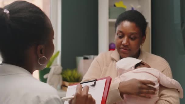 与黑人女儿科医生交谈的年轻黑人妇女抱着新生儿站起来 在医院进行婴儿检查时 在医疗文件中做笔记 — 图库视频影像