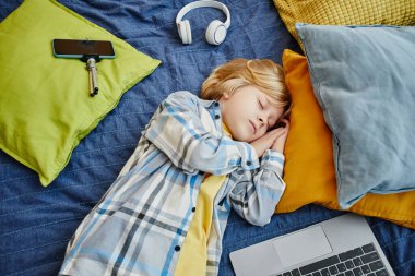 Yastık, dizüstü bilgisayar, akıllı telefon ve kulaklıklar arasında yorgun sarışın okul çocuğu ve blog yazarı yatağında kestiriyor.