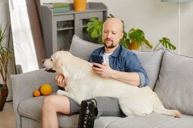 Protez bacaklı, gülümseyen yetişkin bir adamın portresi kanepeye uzanmış mutlu beyaz bir köpek tutuyor.