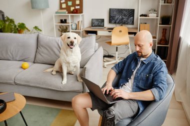 Özürlü yetişkin bir adamın portresi oturma odasında dizüstü bilgisayar kullanarak arka planda beyaz köpek ile, boşluğu kopyala
