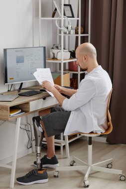Protez bacaklı, evden çalışan ve masa başında doküman okuyan adamın tam boy kenar görüntüsü