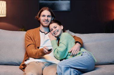 Evlerinde televizyon seyreden ve kucaklaşan gülümseyen genç çiftin ön manzara portresi.