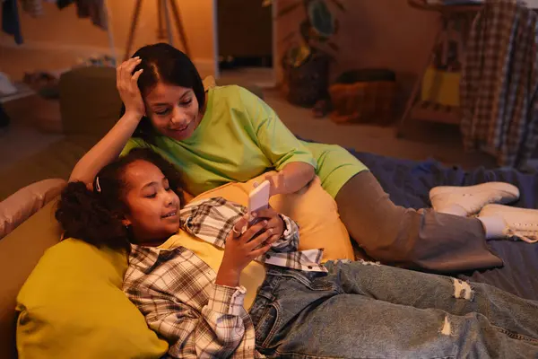 stock image High angle portrait of young mother with daughter lying on bed together and using smartphone in cozy warm light