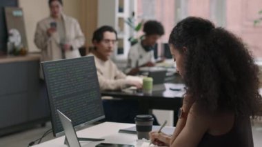Bilgisayar ekranına programlama koduyla bakarken, farklı iş arkadaşlarıyla modern bilişim ofisinde çalışırken, iki ırklı genç bir kadın programcının belden yukarısına bakması.