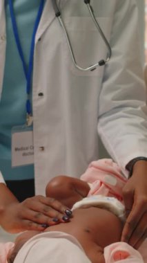 Dikey olarak, Afrikalı Amerikalı kadın pediyatristin bebek kızını muayene ederken ve klinikte tıbbi muayene sırasında annesiyle konuşurken orta eğik pozu.