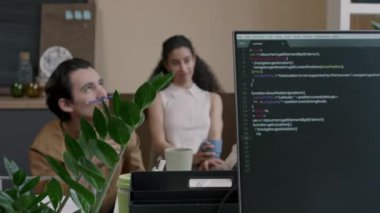 Modern bilişim ofisindeki işler arasında sohbet eden ve dinlenen üç genç çalışan ve önplandaki masaüstü bilgisayarında program kodu olan