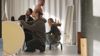 Vücut geliştirme ustası çıplak gövdesiyle ayakta dururken makyaj sanatçısı vücuduna, fotoğrafçısına ve video fotoğrafçısına atölyede reklam çekerken bilgisayar ekranına bakıyor.