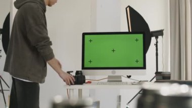 Erkek fotoğrafçının yavaşlaması, profesyonel ışıklandırma ekipmanlarıyla geniş minimalist fotoğraf stüdyosunda yeşil renkli ekranlı DSR kamerayı beyaz masanın yanına yerleştirmesi.