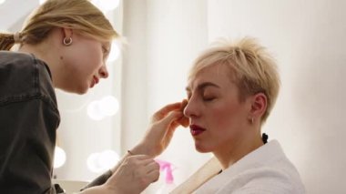Makyajcının beli beyaz, zarif sarışın kadının yüzüne dokunuyor sahne arkasında ampullerle oturuyor ve stüdyo fotoğrafı için hazırlanıyor.