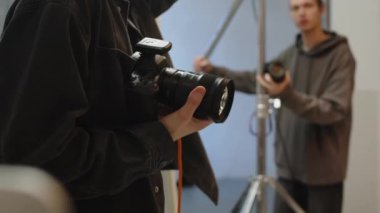 Kafkasyalı erkek fotoğrafçının kamera tutarken ve talimat verirken ve asistanının ışık ekipmanlarını ayarlarken stüdyoda fotoğraf çekerken orta görüntüsü.