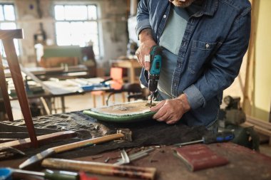 Closeup of senior man repairing old furniture in carpentry workshop and holding electric drilling tool, copy space clipart