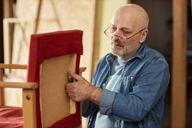 Portrait of experienced senior craftsman restoring old furniture items in workshop clipart