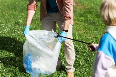 Küçük çocuğun babasıyla birlikte çöpleri topladığı ve plastik şişeyi poşete koyduğu yakın plan.