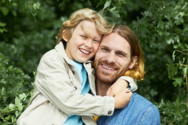 Parkta yeşilliğe sarılan ve kameraya gülümseyen mutlu baba ve oğul portresi.