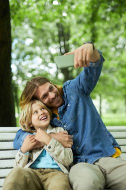 Baba ve oğlunun parkta bankta oturup selfie çekerken çekilmiş portresi.