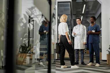 Kendine güvenen, laboratuvar önlüklü olgun bir doktor ve mavi üniformalı asistanı önlerinde duran genç sarışın hastaya danışıyor.