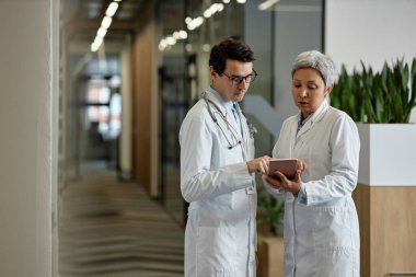 Hastane koridorunda erkek meslektaşlarına tıbbi verileri sunan tabletle kendinden emin kadın pratisyen doktor.