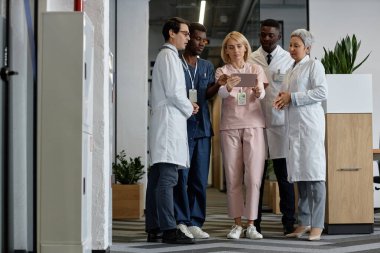 Beş kültürler arası doktor ve üniformalı asistan hastane koridorunda dururken tablet ekranda tıbbi verileri tartışıyorlar.