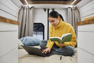 Genç bir kadının dizüstü bilgisayar kullanırken karavanda bacak bacak üstüne atıp yol üstünde çalışırken çekilmiş uzun bir portresi.