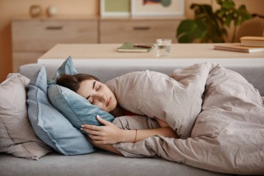 Evdeki kanepede battaniyeye sarılı ve depresyonla boğuşan üzgün genç bir kadının portresi.