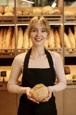 Zanaatkar fırınının arkasında duran taze ekmeği tutan gülümseyen genç bir kadının dik bel portresi.