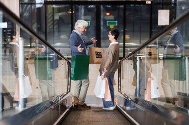 Alışveriş merkezindeki yürüyen merdivende duran alışveriş torbası tutan mutlu çiftin yan görüntüsü.