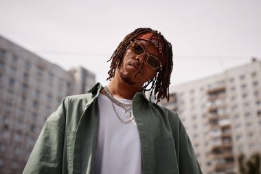 Güneş gözlüklü, şık giyimli, açık havada yüksek binaların önünde kameraya bakan ciddi bir Afrikalı genç adam.