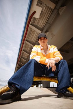 Şapkalı genç bir adam, mavi kot, çizgili kazak ve çapraz ayakkabılı. Köprünün altında mavi gökyüzüne karşı otururken kameraya bakıyor.