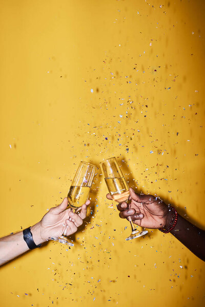 Close up of two people clinking champagne glasses against vibrant yellow background with confetti pop