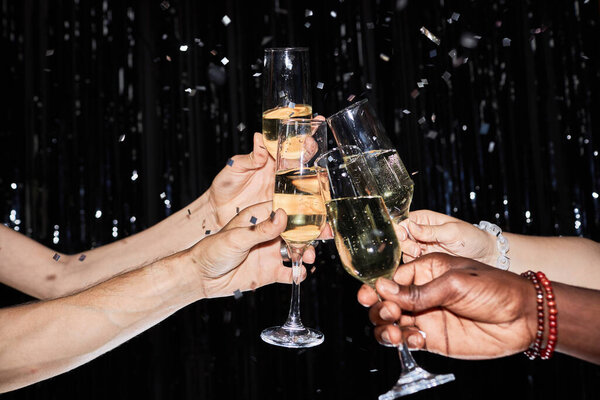 Closeup of hands toasting with champagne glasses at party against glittering background with confetti