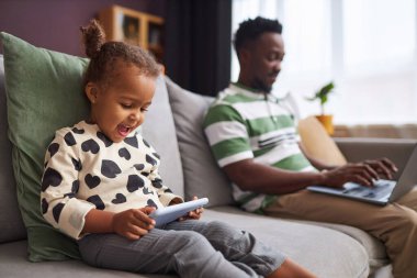 Küçük bir kızın arka planda çalışan babasıyla birlikte akıllı telefon kullandığı yan görünüm portresi.
